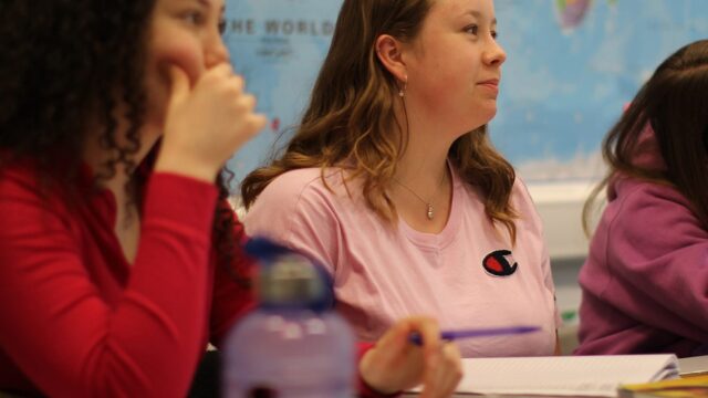 Students sat at classroom tables looking towards the front. There is a world map in the background.