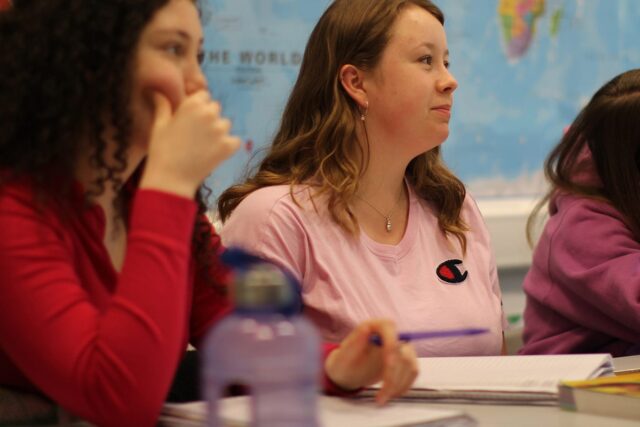 Students sat at classroom tables looking towards the front. There is a world map in the background.