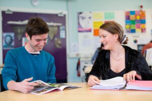 Two students sat at a classroom table flipping through books and folders.