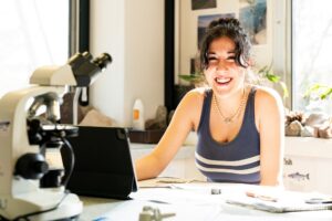 A student sits at a lab table and smiles at the camera. She has a tablet and a microscope in front of her.