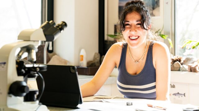 A student sat in a laboratory with a tablet and a microscope in front of her.