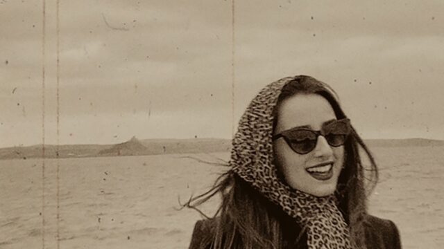 A sepia toned image of a woman in a headscarf and sunglasses with the sea and St Michaels Mount in the background.