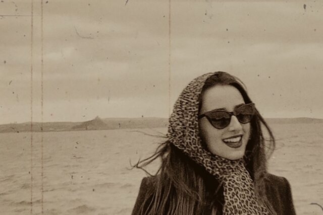 A sepia toned image of a woman in a headscarf and sunglasses with the sea and St Michaels Mount in the background.