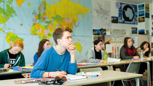 A student sits at a classroom table with his hand to his chin. Students are sat behind.