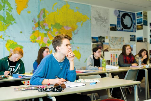 A student sits at a classroom table with his hand to his chin. Students are sat behind.