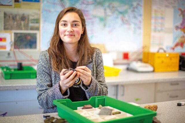 A student smiles at the camera holding a rock. There is a tray of rocks in front of them.
