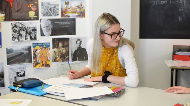 A student working at a classroom table with notebooks and pens. There are posters on the wall next to her.