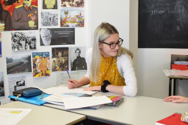A student working at a classroom table with notebooks and pens. There are posters on the wall next to her.