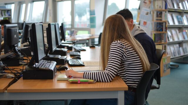 Two students sit in a library at computers.