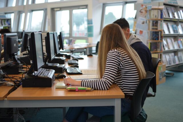 Two students sit in a library at computers.