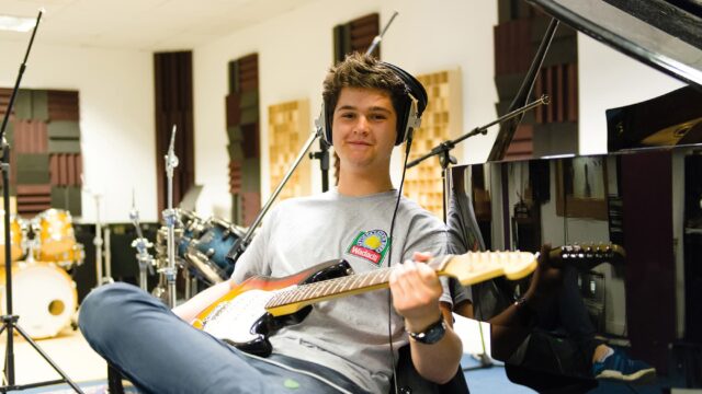 A student sat on a chair with a guitar and wearing headphones. There are lots of instruments in the room.