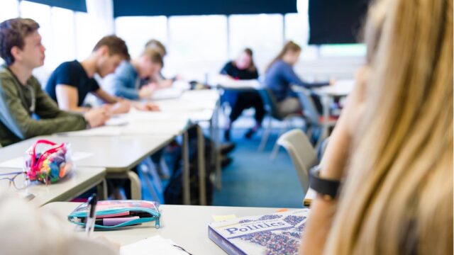 Looking over the shoulder of a student towards a classroom table that has a book labelled 'Politics' on it.