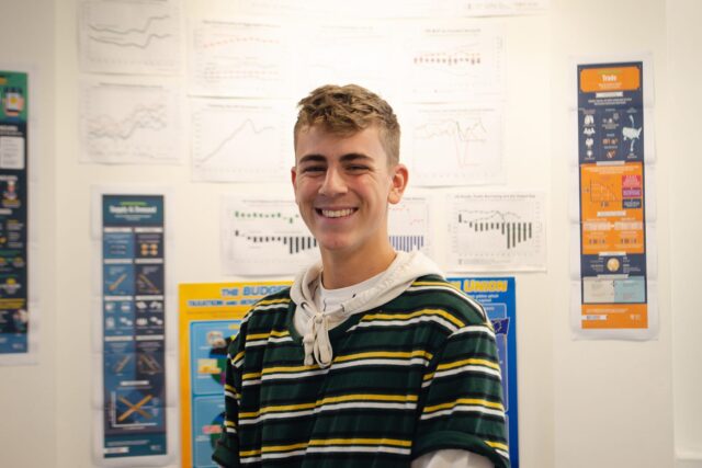 A student smiling. In the background there is a wall covered in posters.