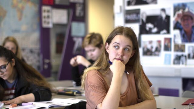 A student sat at a classroom table with her hand under her chin and a pencil in her hand. There are students in the background working at classroom tables.