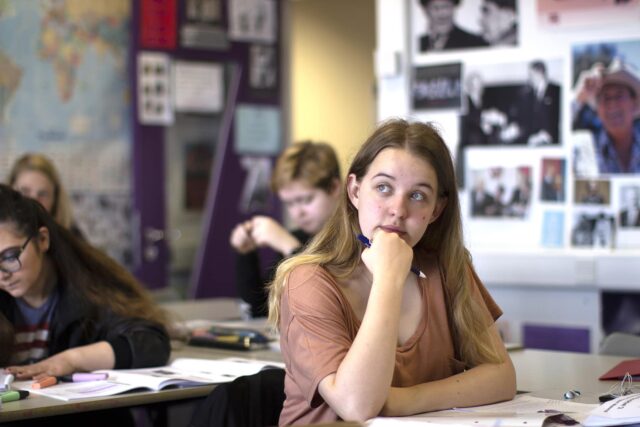 A student sat at a classroom table with her hand under her chin and a pencil in her hand. There are students in the background working at classroom tables.