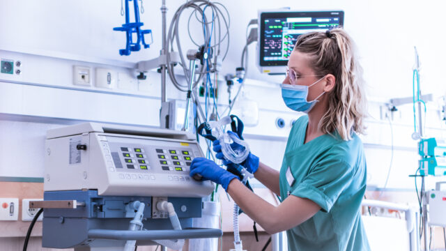 Nurse, wearing a surgical mask, checks the settings of a mechanical ventilation machine, which is seen in the foreground. Therapy used for lung breathing, in intensive care.