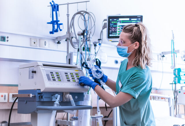 Nurse, wearing a surgical mask, checks the settings of a mechanical ventilation machine, which is seen in the foreground. Therapy used for lung breathing, in intensive care.