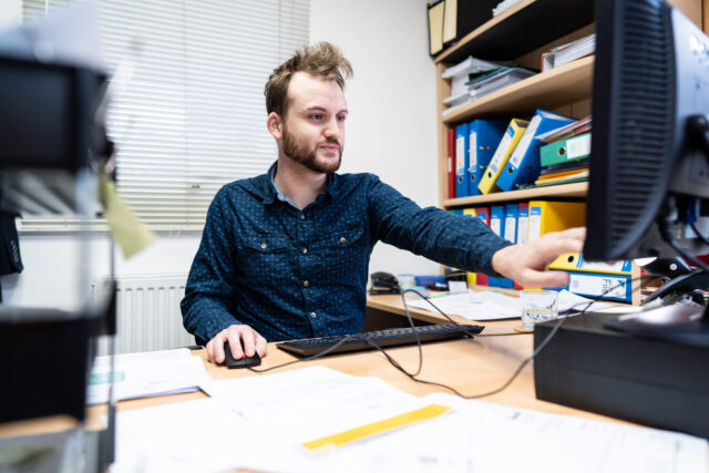A man in a blue shirt sits at a desk and points towards the computer in front of him.