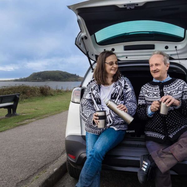 A couple perch on the boot of a vehicle. One holds a cup while the other pours from a flask.
