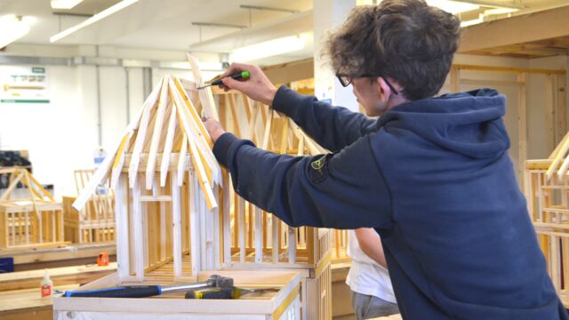 A student is working on a miniature wooden structure of a building.