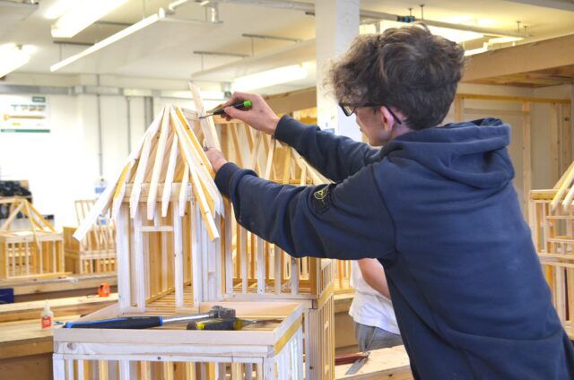 A student is working on a miniature wooden structure of a building.