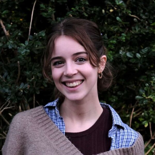 Alex is stood in front of greenery and is smiling.