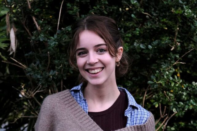 Alex is stood in front of greenery and is smiling.