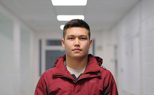 Ali is stood in a corridor and is wearing a red coat.