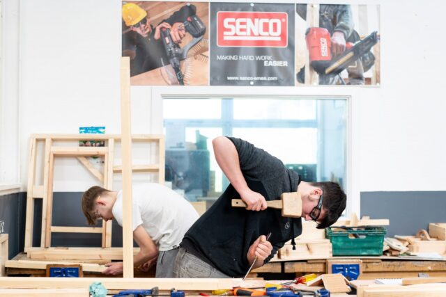 Two students are using a hammer and chisel on wood.