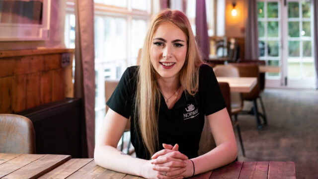 An apprentice sits at a table in a pub with their hands folded on the table.