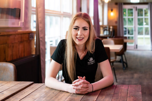 An apprentice sits at a table in a pub with their hands folded on the table.