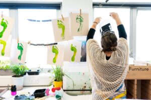 A tutor hanging prints of green footprints to a washing line within an art classroom.