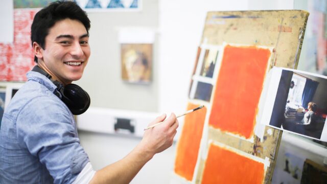 A student with a paintbrush in hand and headphones around his neck. He is painting large squares of orange on an easel.