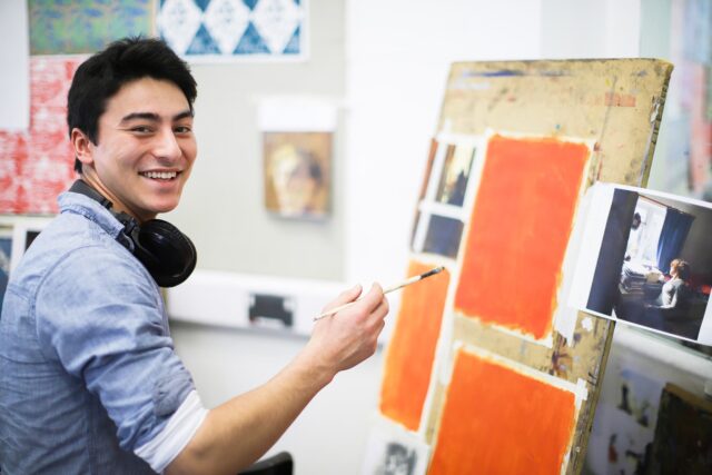 A student with a paintbrush in hand and headphones around his neck. He is painting large squares of orange on an easel.