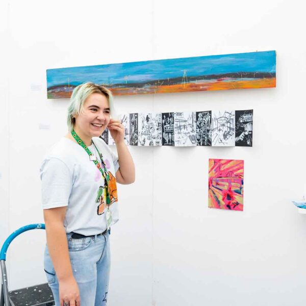 A student stands in the centre of a white exhibition space. There are a few pieces if art work on the walls and a step ladder too.
