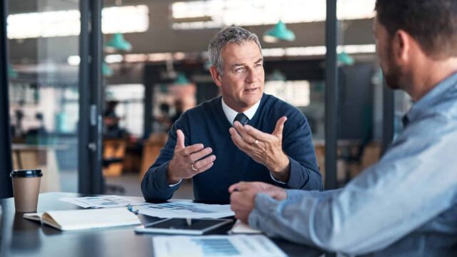 Two men sit at an office table and talk while there is paperwork in front of them.