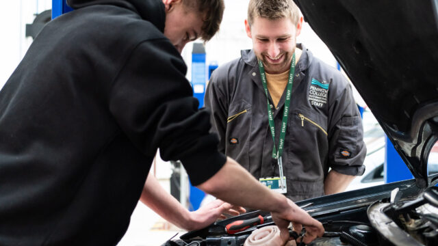 A tutor watches as a student looks under the bonnet of a car.