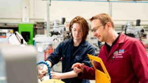 An engineering tutor looks at equipment with a student. They are both wearing overalls.