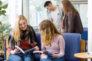 Two students sit on a sofa and look at books together.