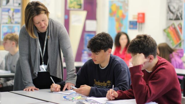 Students working in a classroom while a tutor stands over them to help.