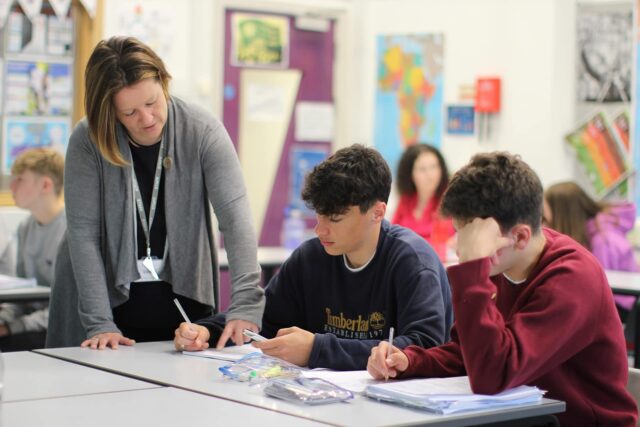 Students working in a classroom while a tutor stands over them to help.