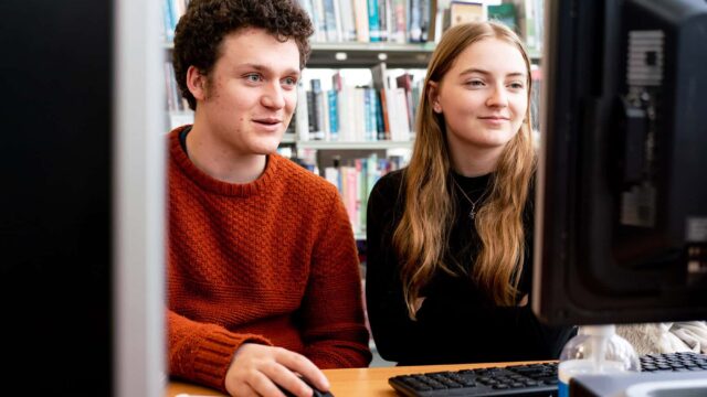Two students sit at a computer together within a College library.