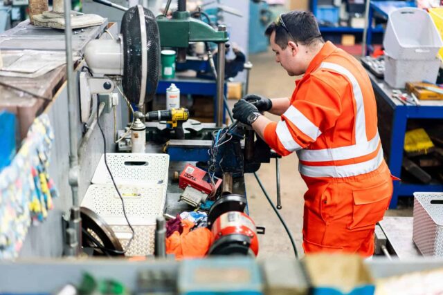 An apprentice in orange overalls works on a piece of machinery.