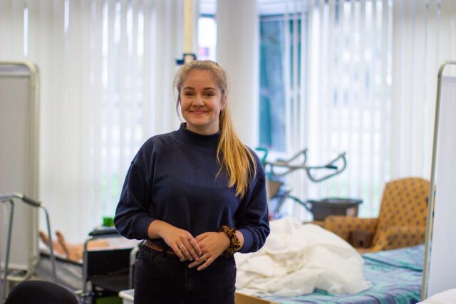 A student stands in front of a hospital bed and smiles.