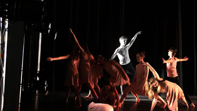 A group of performers on the stage. They are all wearing simple outfits with dramatic lighting.