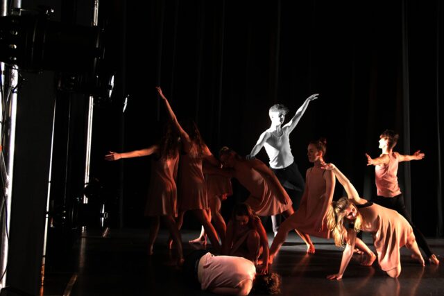 A group of performers on the stage. They are all wearing simple outfits with dramatic lighting.