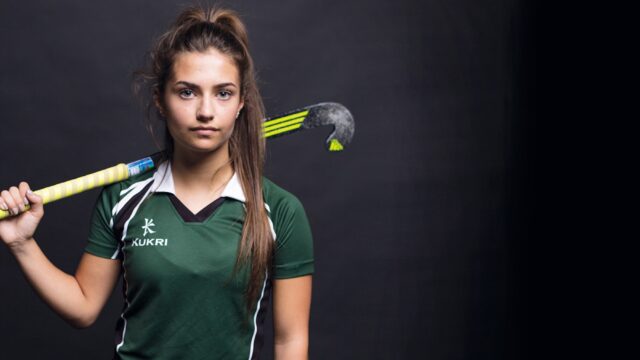 A student hockey player in a green uniform with a hockey stick up on her shoulder.
