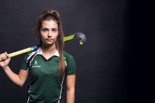 A student hockey player in a green uniform with a hockey stick up on her shoulder.