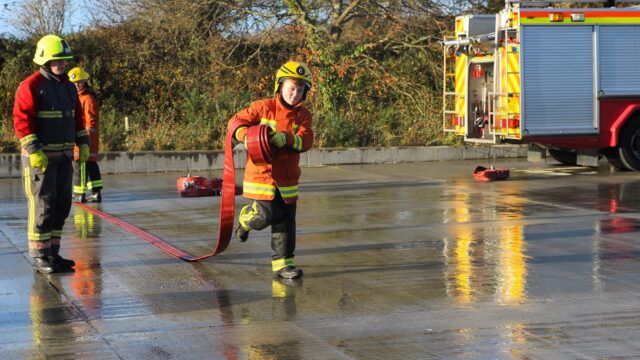 Students dressed as firefighters laying out a hose with a fire engine in the background.