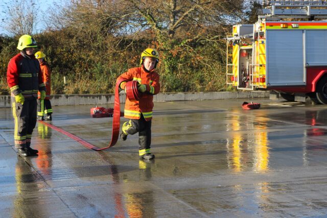 Students dressed as firefighters laying out a hose with a fire engine in the background.